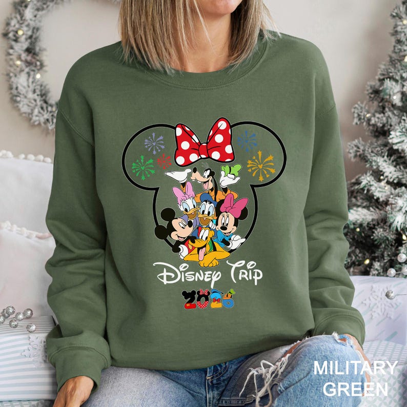 May include: An olive green sweatshirt featuring a large graphic of Mickey Mouse and friends. The design includes a red polka dot bow, fireworks, and the words "Disney Trip". The sweatshirt is a comfortable, casual style.