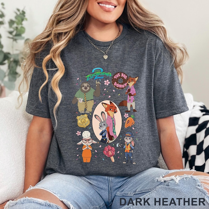 K&ouml;nnte beinhalten: Ein dunkelgrau meliertes T-Shirt mit einem Zootopia-Grafikdruck. Das Design zeigt Charaktere wie Judy Hopps und Nick Wilde, eine Polizeimarke, einen Donut, eine Karotte und ein pfotenf&ouml;rmiges Eis am Stiel, umgeben von kleinen rosa Bl&uuml;tenbl&auml;ttern.