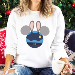May include: White sweatshirt featuring a cartoon design of a police officer bunny. The design includes a blue uniform, a yellow badge, and gray ears. The sweatshirt is a crew neck style.