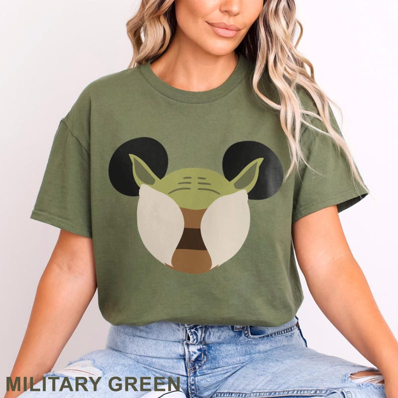 May include: Military green t-shirt featuring a graphic of Yoda's face with Mickey Mouse ears. The design includes shades of green, brown, and white. The text "MILITARY GREEN" is at the bottom of the image.