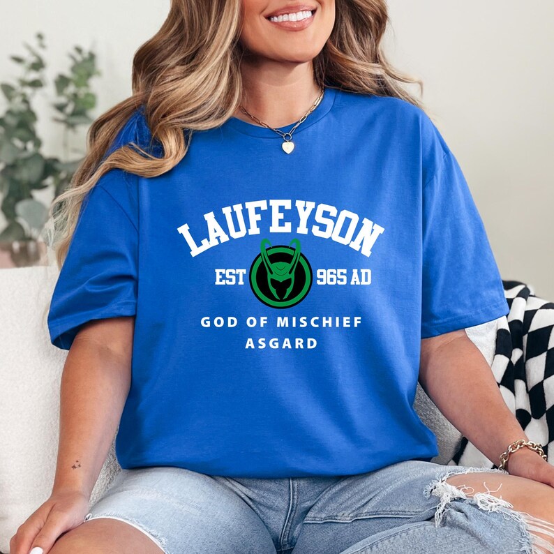 May include: Royal blue t-shirt with the word "LAUFEYSON" in white arched text. Below, a green and black emblem is flanked by "EST" and "965 AD". The text "GOD OF MISCHIEF ASGARD" is printed below the emblem.