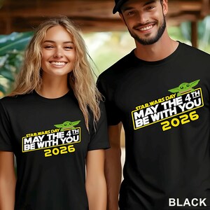 May include: Black t-shirts featuring the text "STAR WARS DAY MAY THE 4TH BE WITH YOU 2026" in yellow and white, with a green cartoon character. The shirts are worn by two people.