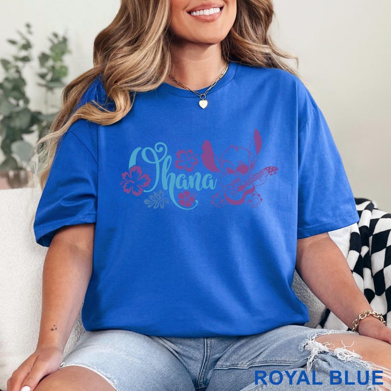 Op de afbeelding: Een koningsblauw t-shirt met korte mouwen en een grafische print in lichtblauw en magenta. Het ontwerp bevat het woord Ohana in cursief schrift, gestileerde hibiscusbloemen en een silhouet van het personage Stitch die ukelele speelt.