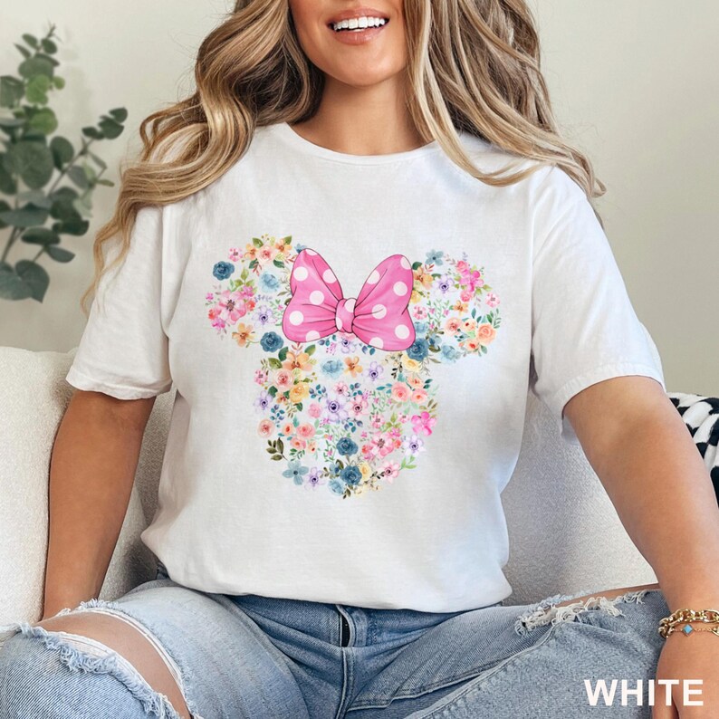 May include: White t-shirt featuring a floral Minnie Mouse design. The design includes a pink polka-dot bow and a floral arrangement in the shape of Minnie's head. The shirt is made of a soft material and is suitable for casual wear.