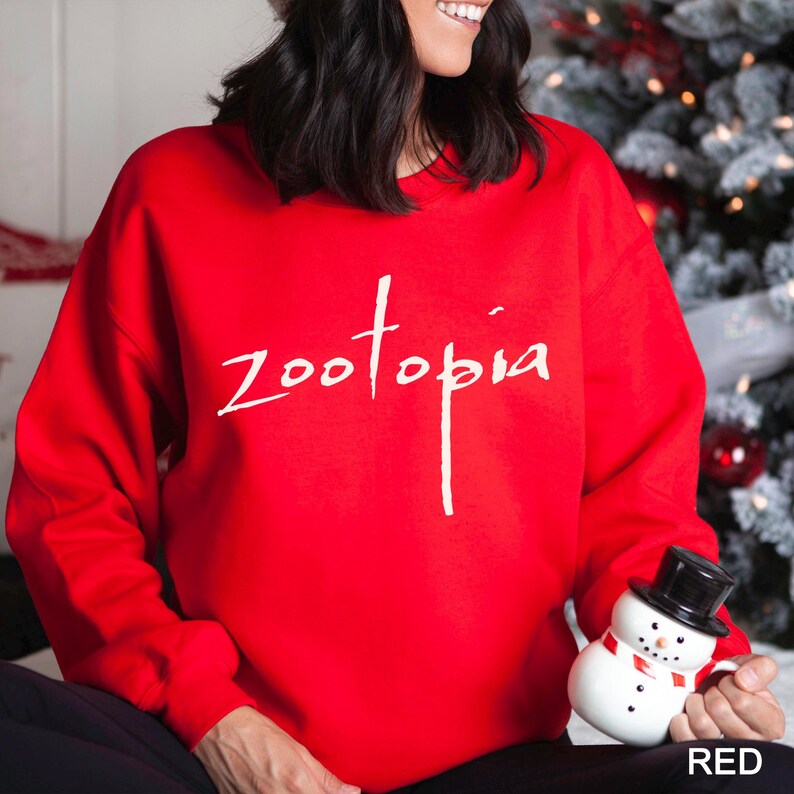May include: A bright red sweatshirt with the word "Zootopia" in white script. The sweatshirt is a solid color and has a crew neck. A small snowman figurine is held in the person's lap. The word "RED" is in the bottom right corner.