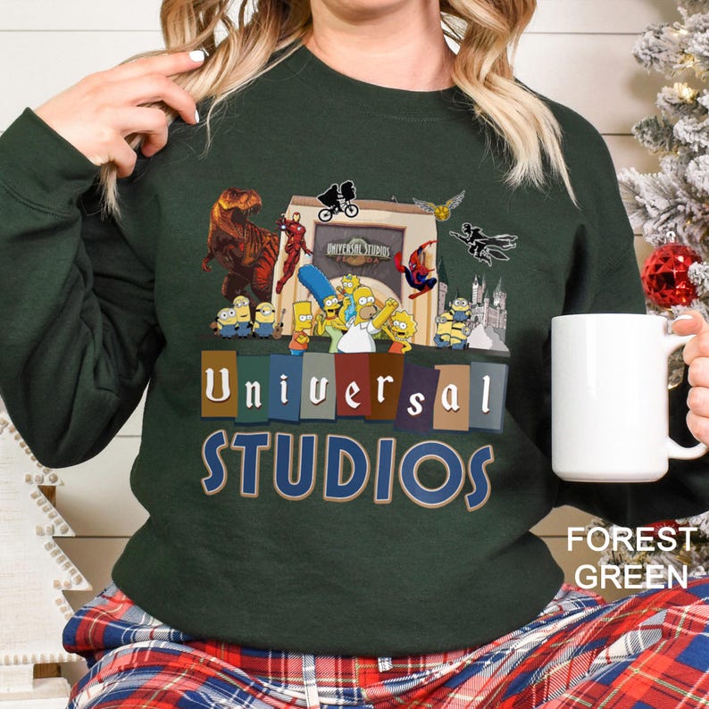 May include: Forest green sweatshirt featuring a Universal Studios graphic with various cartoon characters and the text "Universal Studios". The sweatshirt is paired with plaid pajama pants and a white mug.
