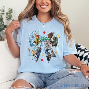 Op de afbeelding: Lichtblauw T-shirt met een groot strikdesign met personages uit de film "Zootopia". De strik is gevuld met afbeeldingen van de personages en kleine decoratieve elementen. De tekst "LIGHT BLUE" staat onderaan.