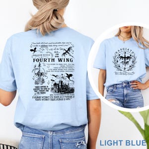 May include: Light blue t-shirts with graphic designs. One shirt features the text "Fourth Wing" with dragon and castle illustrations. The other shirt displays "Basgiath War College" with a dragon and moon phases. Both shirts are made of soft material.