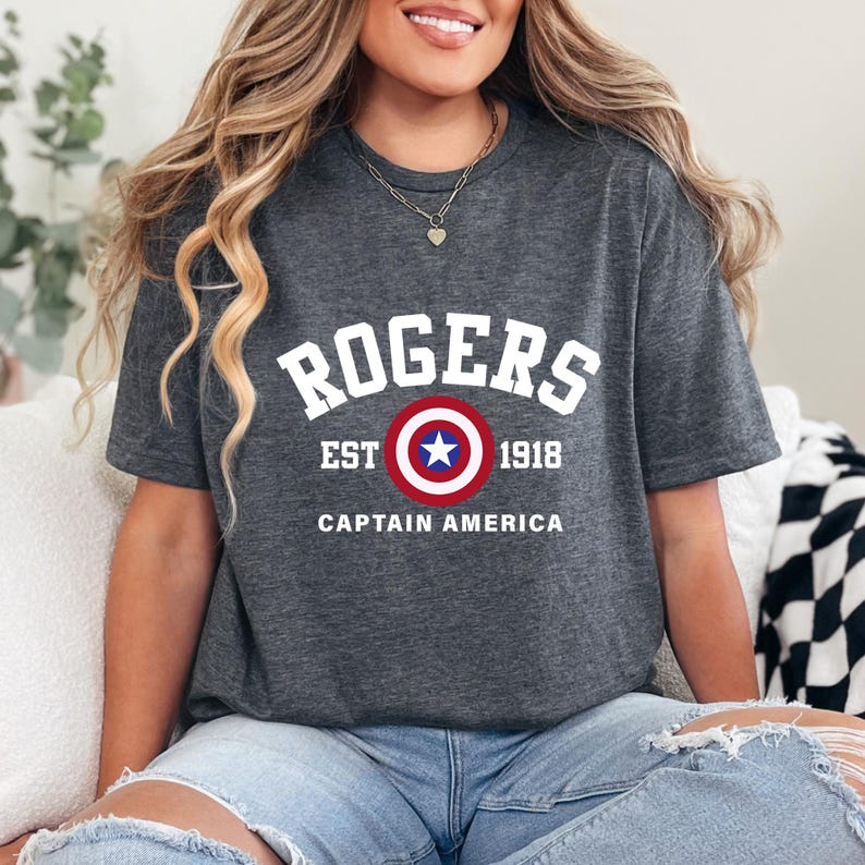 May include: Dark gray t-shirt featuring the name "ROGERS" in white block letters above a shield design with a red, white, and blue color scheme. The text "EST 1918" and "CAPTAIN AMERICA" are also printed on the shirt.