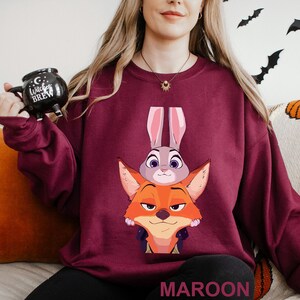 May include: Maroon sweatshirt featuring a cartoon illustration of a fox and a bunny. The fox is orange with a stern expression, and the bunny is gray. The word "MAROON" is printed at the bottom. The sweatshirt is a solid maroon color.