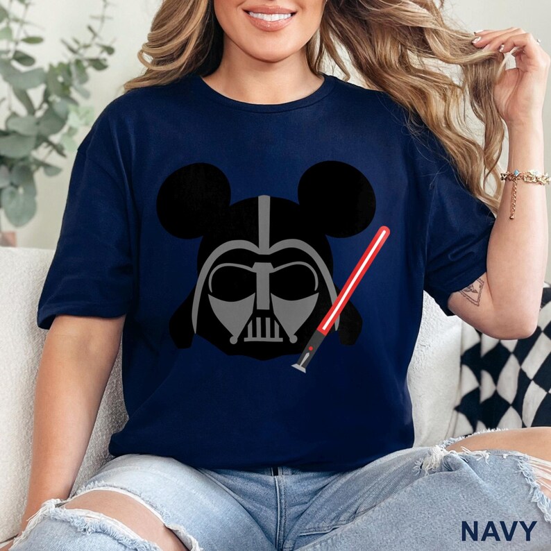 May include: Navy blue t-shirt featuring a graphic of Darth Vader's helmet with Mickey Mouse ears and a red lightsaber. The shirt's design is a fusion of Star Wars and Disney themes. The word "NAVY" is printed at the bottom.