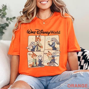 May include: Bright orange t-shirt featuring a Walt Disney World graphic. The design includes four panels with cartoon images of a fox and a rabbit. The text "Walt Disney World" is above the panels, and "ORANGE" is at the bottom.