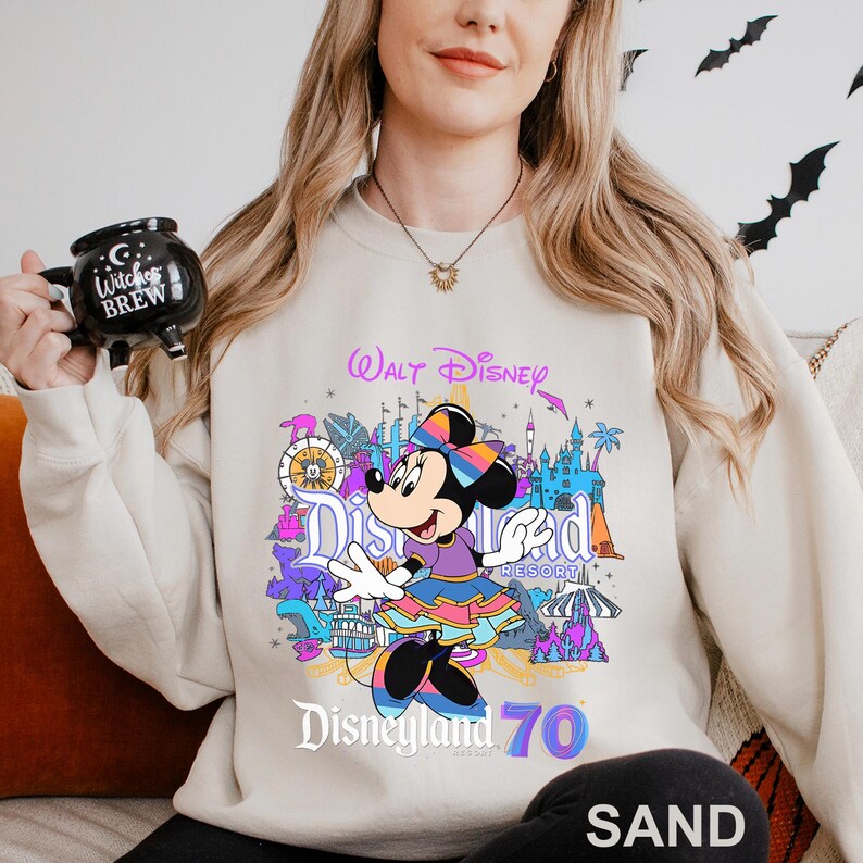 May include: Sand-colored sweatshirt featuring a colorful graphic of Minnie Mouse in front of Disneyland Resort. The design includes the words "Walt Disney," "Disneyland," and "70." The sweatshirt is a comfortable, casual style.