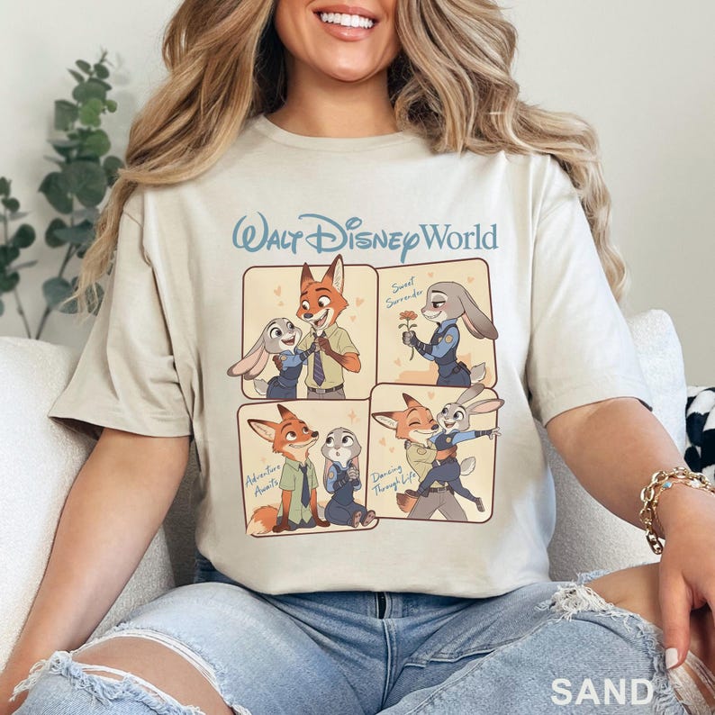 K&ouml;nnte beinhalten: Sandfarbenes T-Shirt mit einem Walt Disney World-Logo und einer viergeteilten Illustration der Zootopia-Charaktere Nick Wilde und Judy Hopps. Die Panels zeigen die Charaktere in verschiedenen Posen, mit dem Text "Sweet Surrender", "Adventure Awaits" und "Dancing Through Life".