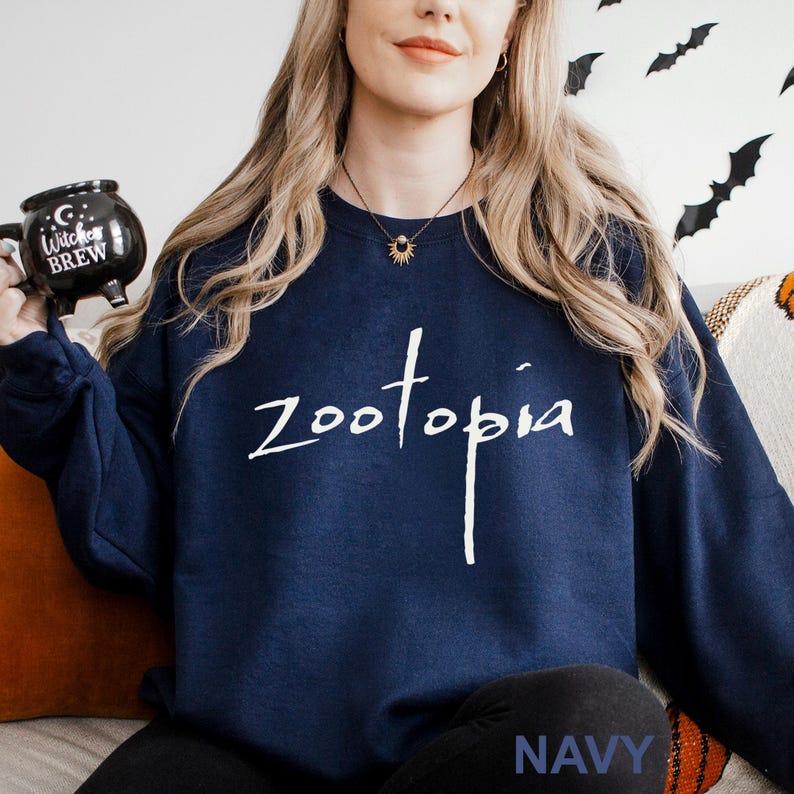May include: Navy blue sweatshirt with the word "zootopia" in white script. The word "NAVY" is printed in white at the bottom. A black mug with the words "Witch's Brew" is held in the left hand.