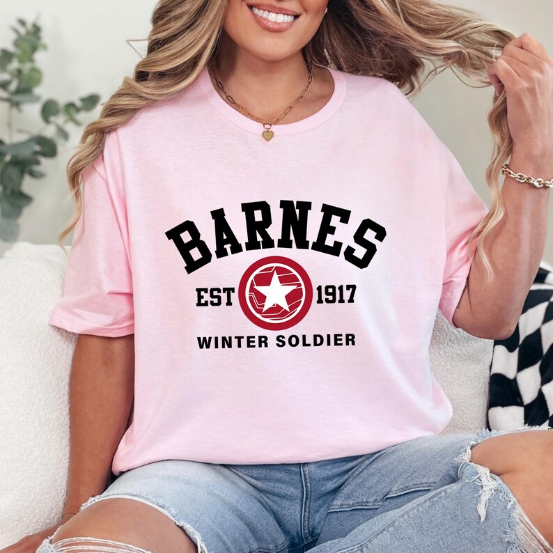 May include: A light pink t-shirt with the word "BARNES" in bold black letters. Below the word is a red circle with a white star and the text "EST 1917 WINTER SOLDIER". The shirt is a crew neck style.
