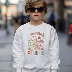 Pode incluir: Uma camisola branca com o texto "UNIVERSAL STUDIOS" num design colorido em arco. O design inclui v&aacute;rios personagens e &iacute;cones de desenhos animados. A camisola &eacute; usada por uma pessoa que usa &oacute;culos de sol.