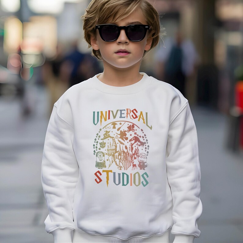 Pode incluir: Uma camisola branca com o texto "UNIVERSAL STUDIOS" num design colorido em arco. O design inclui v&aacute;rios personagens e &iacute;cones de desenhos animados. A camisola &eacute; usada por uma pessoa que usa &oacute;culos de sol.