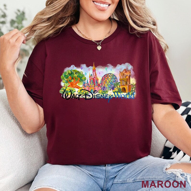 May include: A maroon short-sleeve t-shirt featuring a colorful graphic print of Walt Disney World landmarks, including a tree, a castle, a geodesic sphere, and a tower, with the text "Walt Disney World" printed in a dark blue script font.