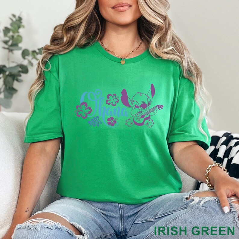 Op de afbeelding: Een felgroen t-shirt met korte mouwen en een paarse grafische print van een stripfiguur die ukelele speelt, omringd door hibiscusbloemen en het woord Ohana in een gestileerd lettertype, gecentreerd op de borst van het kledingstuk.