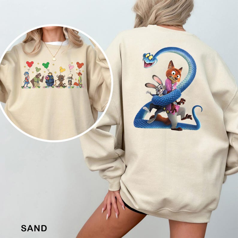 May include: A sand-colored crewneck sweatshirt featuring a large graphic on the back of a blue snake coiled around a fox and a rabbit. The front displays a smaller graphic of six animated characters holding colorful balloons in a horizontal row.