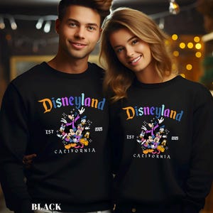 Puede incluir: Sudaderas negras con el logo de Disneyland en naranja y azul, con Mickey Mouse y sus amigos. El texto "EST 2025 CALIFORNIA" est&aacute; debajo del gr&aacute;fico. La palabra "BLACK" est&aacute; en la parte inferior de la imagen.