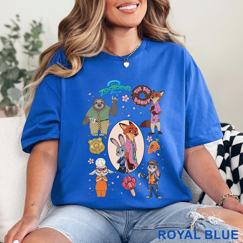 K&ouml;nnte beinhalten: Ein k&ouml;nigsblaues T-Shirt mit einem Zootopia-Grafikdruck, der Charaktere wie Flash, Nick Wilde, Judy Hopps und Bellwether zeigt. Das Design enth&auml;lt den Text Zootopia und The Big Donut sowie Symbole wie ein Abzeichen, eine Karotte und ein pfotenf&ouml;rmiges Eis.