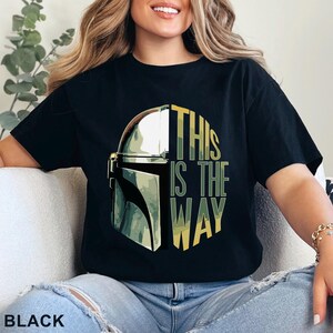 Pu&ograve; includere: Una t-shirt nera a maniche corte con una stampa grafica di un casco metallico sul lato sinistro e il testo "THIS IS THE WAY" in lettere gialle e verdi sul lato destro, su uno sfondo scuro.