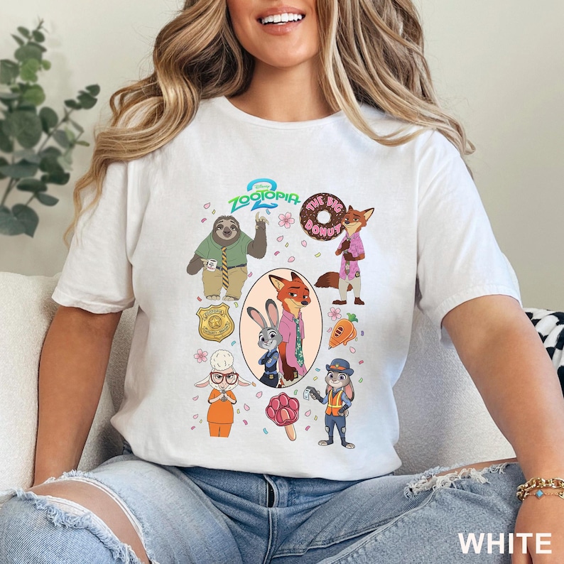 K&ouml;nnte beinhalten: Ein wei&szlig;es Kurzarm-T-Shirt mit einem farbenfrohen Zootopia 2 Grafikdruck. Das Design zeigt Charaktere wie Judy Hopps, Nick Wilde und Flash sowie Symbole wie einen Donut, eine Karotte und eine Polizeimarke, die zentral angeordnet sind.
