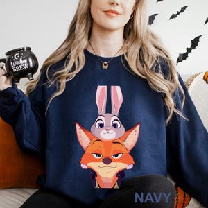 May include: Navy blue sweatshirt featuring a cartoon illustration of a fox and a bunny. The fox is orange and the bunny is gray. The word "NAVY" is printed in white at the bottom of the sweatshirt. The sweatshirt is a crew neck style.