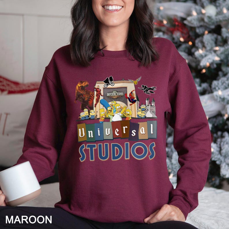 May include: Maroon sweatshirt featuring a colorful graphic of Universal Studios characters and logo. The design includes characters from The Simpsons, Minions, and other popular franchises. The text "Universal Studios" is prominently displayed.