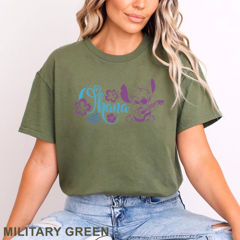 Op de afbeelding: Een militair groen t-shirt met korte mouwen en een grafische print in lichtblauw en paars. Het ontwerp bevat het woord Ohana in een cursief lettertype, diverse hibiscusbloemen en een stripfiguur die een klein snaarinstrument bespeelt.