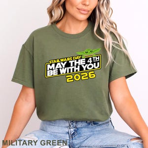 May include: Military green t-shirt featuring the text "STAR WARS DAY MAY THE 4TH BE WITH YOU 2026" in yellow and white, with a green cartoon character graphic. The shirt is a crew neck style.