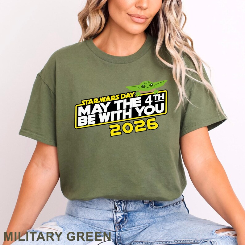 May include: Military green t-shirt featuring the text "STAR WARS DAY MAY THE 4TH BE WITH YOU 2026" in yellow and white, with a green cartoon character graphic. The shirt is a crew neck style.