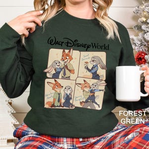 May include: Forest green sweatshirt featuring a four-panel graphic of Nick Wilde and Judy Hopps from Zootopia. The top panel reads "Walt Disney World" and the bottom right panel reads "Dancing Through Life". A white mug is held in the hand.