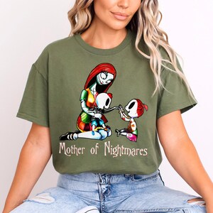Puede incluir: Camiseta verde oliva con un gr&aacute;fico colorido de un personaje con cabello rojo y costuras, sosteniendo una mu&ntilde;eca esqueleto e interactuando con un ni&ntilde;o. El texto "Mother of Nightmares" est&aacute; impreso debajo.