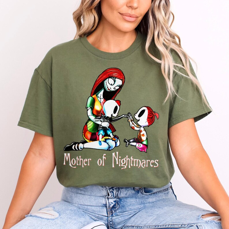 Puede incluir: Camiseta verde oliva con un gr&aacute;fico colorido de un personaje con cabello rojo y costuras, sosteniendo una mu&ntilde;eca esqueleto e interactuando con un ni&ntilde;o. El texto "Mother of Nightmares" est&aacute; impreso debajo.
