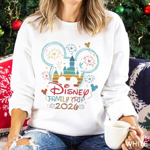 May include: White sweatshirt with a colorful graphic design featuring a castle, fireworks, and Mickey Mouse ears. The text "Disney Family Trip 2026" is also displayed. The sweatshirt is a crew neck style.