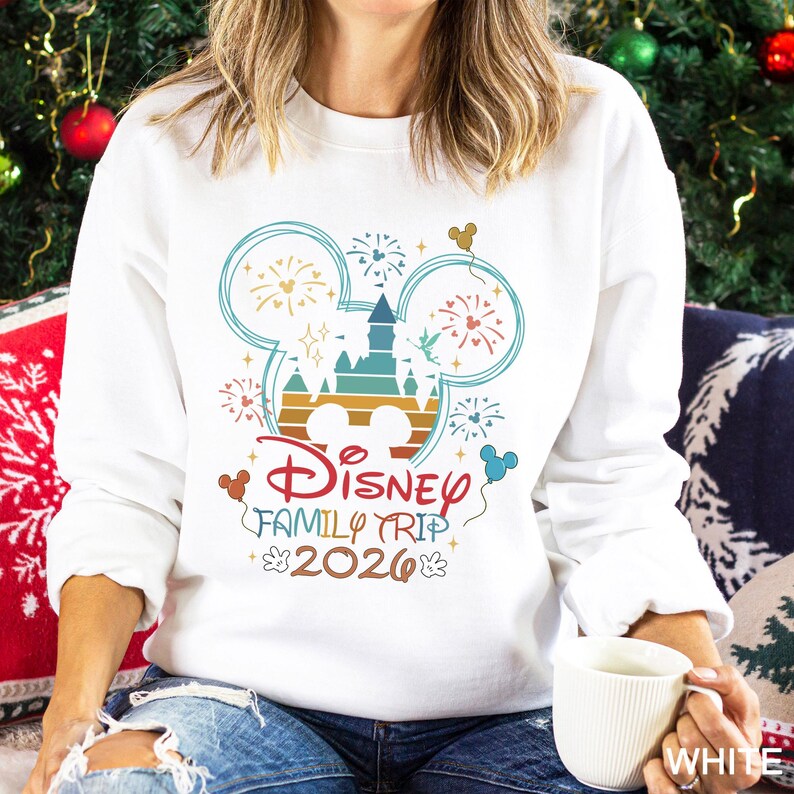May include: White sweatshirt with a colorful graphic design featuring a castle, fireworks, and Mickey Mouse ears. The text "Disney Family Trip 2026" is also displayed. The sweatshirt is a crew neck style.