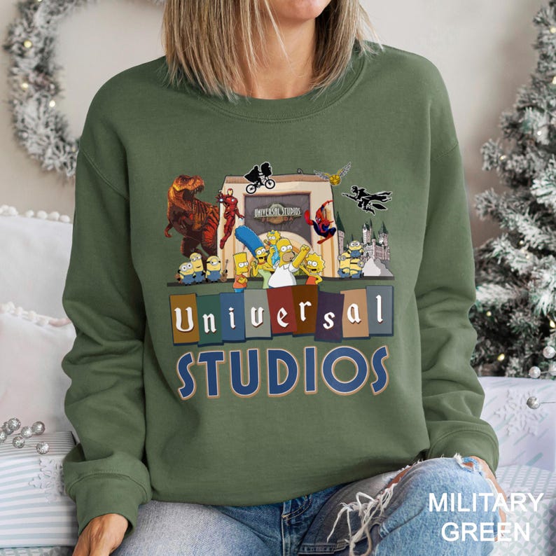 May include: Military green sweatshirt featuring a colorful graphic of Universal Studios characters, including dinosaurs, superheroes, and cartoon figures. The words "Universal Studios" are displayed in a block letter design.