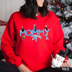 K&ouml;nnte beinhalten: Ein leuchtend rotes Langarm-Sweatshirt mit dem Wort MOMMY in einer blauen, eisigen Schriftart. Die Grafik enth&auml;lt verschiedene animierte Figuren aus der Frozen-Filmreihe, die um die Buchstaben herum angeordnet sind, darunter ein Rentier und ein Schneemann.
