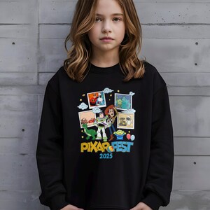Może przedstawiać: Czarna bluza z kolorową grafiką postaci z film&oacute;w Pixar, w tym Buzz Astral i Woody, z napisem "PIXAR FEST 2025". Projekt zawiera obrazy innych postaci i element&oacute;w z film&oacute;w Pixar.