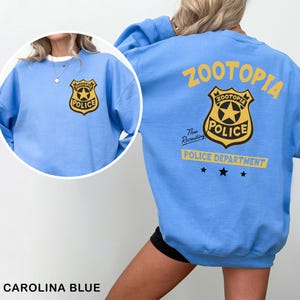 May include: Carolina blue sweatshirt featuring a Zootopia Police badge on the front and back. The back of the sweatshirt has the word "Zootopia" in yellow, a police badge, and the words "Police Department".
