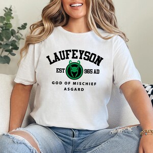 May include: White t-shirt with the word "LAUFEYSON" in black, arched above a green and black emblem. Below the emblem are the words "EST 965 AD" and "GOD OF MISCHIEF ASGARD" in black. The shirt is worn by a person.