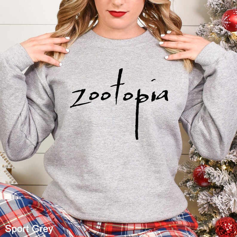May include: A light gray sweatshirt with the word "Zootopia" in black script across the chest. The sweatshirt is a crew neck style. The bottom of the image shows a plaid pattern.