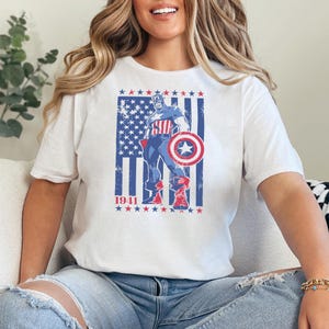May include: White t-shirt featuring a graphic of Captain America in a patriotic design. The design includes the American flag, stars, and the year "1941". The superhero is depicted holding his shield, with a vintage, distressed aesthetic.