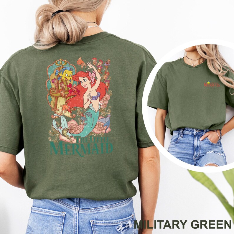 May include: An olive green t-shirt featuring a large graphic on the back of Ariel and Flounder from The Little Mermaid surrounded by floral elements and the text The Little Mermaid. The front chest area displays a small graphic of Flounder.