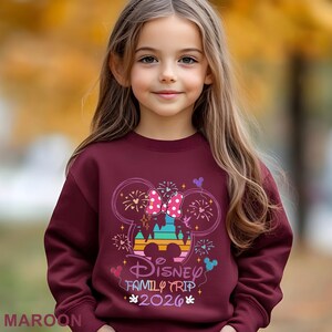 May include: A maroon sweatshirt featuring a colorful graphic of Minnie Mouse's head with a bow, fireworks, and the text "Disney Family Trip 2026". The sweatshirt is worn by a young girl.