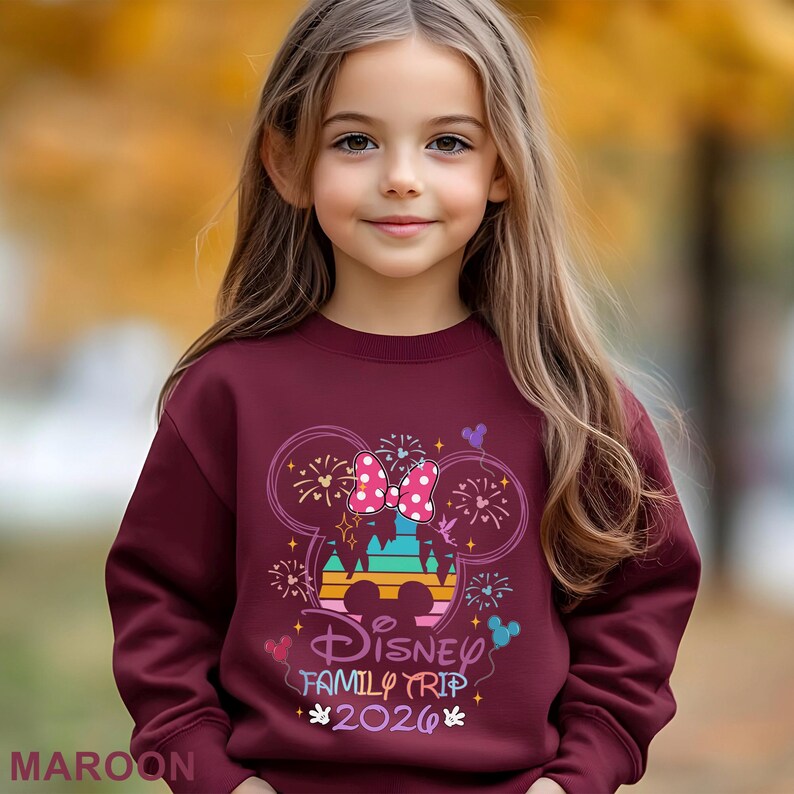 May include: A maroon sweatshirt featuring a colorful graphic of Minnie Mouse's head with a bow, fireworks, and the text "Disney Family Trip 2026". The sweatshirt is worn by a young girl.