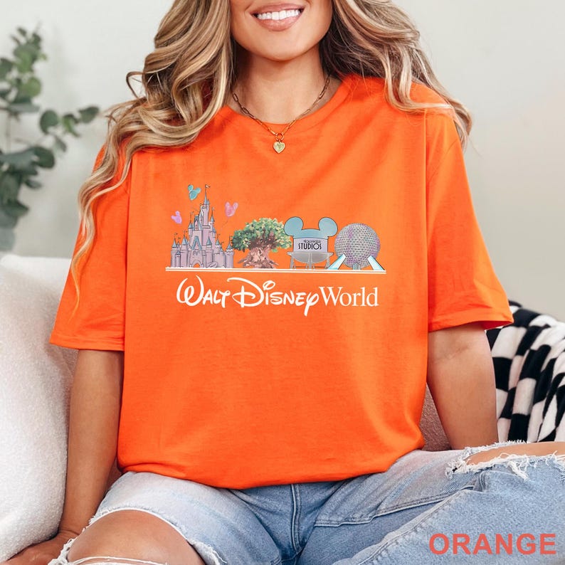 Op de afbeelding: Een feloranje T-shirt met korte mouwen en een afbeelding van Walt Disney World-bezienswaardigheden, waaronder een kasteel, een boom, een Hollywood Studios-bord en een geodetische bol, met de tekst Walt Disney World in het wit eronder.